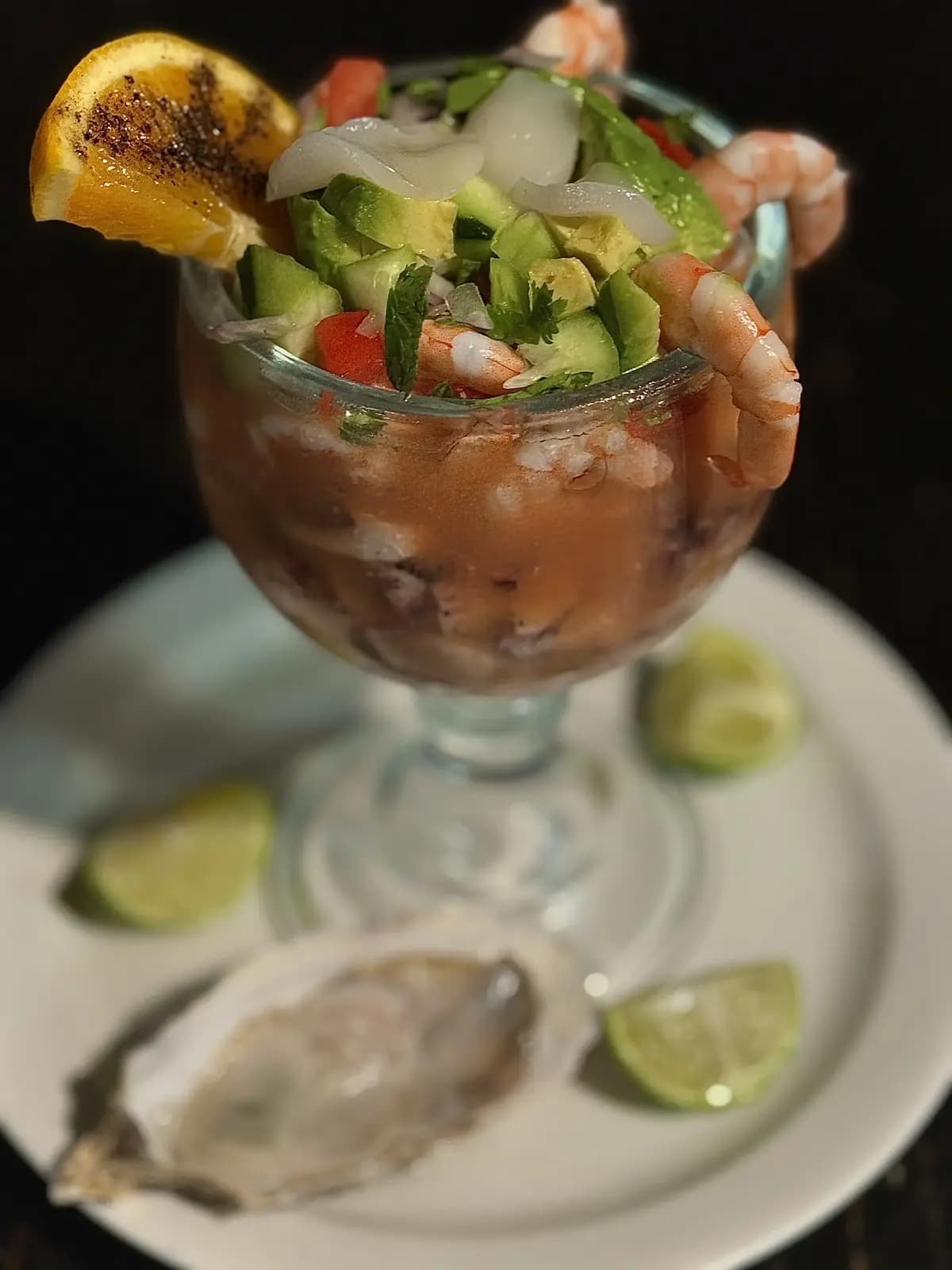 Glass cocktail cup packed and overflowing with shrimp, cucumber, onions, tomato, and scallops. It sits on a plate with lime and an oyster.