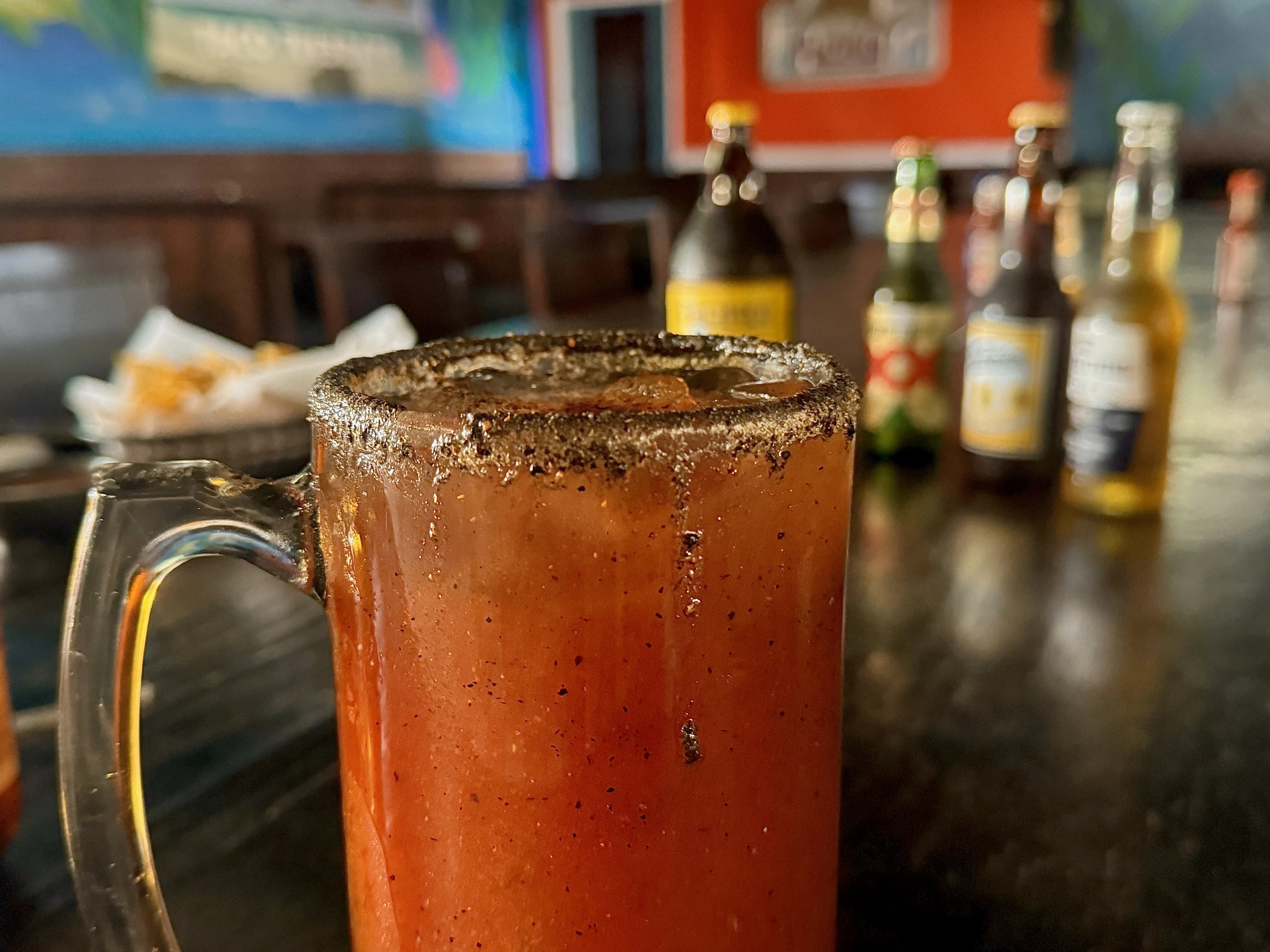 Ice cold red/orange michelada served in a large glass Tankard cup. The cup is seating with precipitation and a resturant dining area in the background with chips and beer.