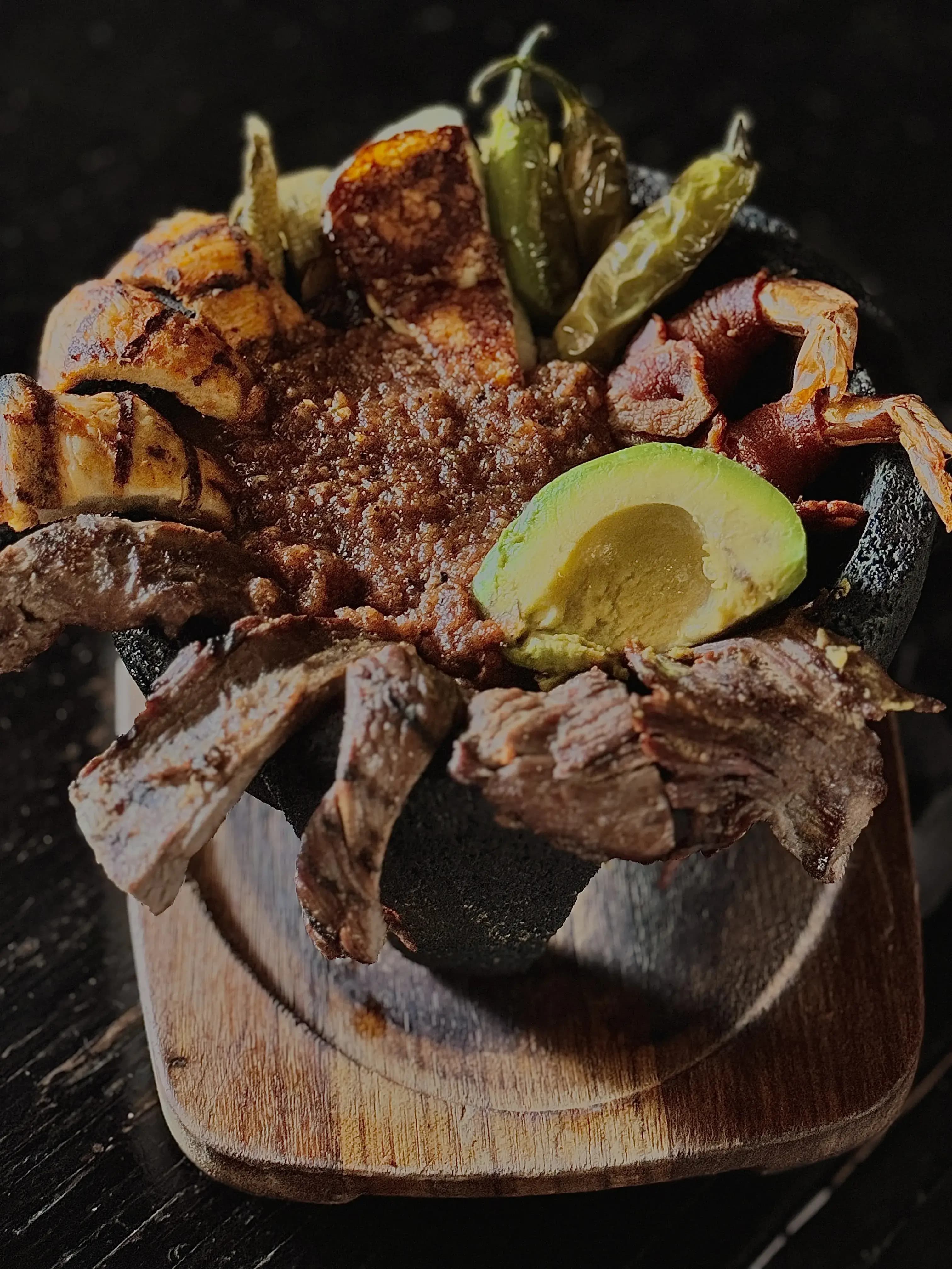 Stone and rock bowl filled with a sizzling sauce that from which emerges grilled chicken, carne asada, nopal, hot peppers, bacon-wrapped shrimp, and avocado.